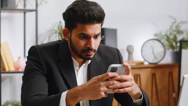 Indian businessman in suit use smartphone typing browsing, loses becoming surprised sudden lottery results, bad news, fortune loss fail at home office desk workplace. Bearded freelancer worker man