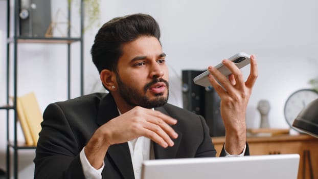 Indian businessman working, having mobile phone loudspeaker talk at home office desk with laptop. Bearded professional freelance man holding smartphone using messenger chat apps. Employment occupation