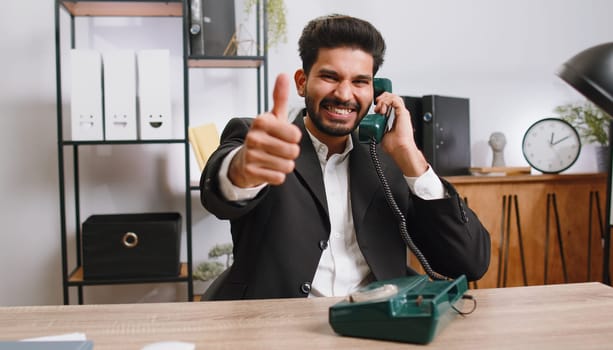 Hey you, call me back. Bearded indian businessman talking on wired retro vintage telephone of 80s at home office workplace desk. Male hindu in formal suit. Remote distant online working on laptop