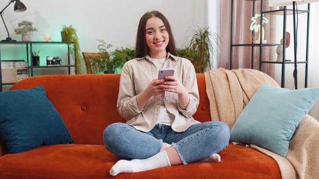Portrait of young woman sitting on sofa uses mobile phone smiling at home living room apartment. Girl texting share messages content on smartphone social media applications online watching relax movie