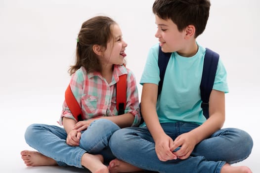 Portrait of lovely brother and sister, elementary age school kids with backpacks - teen boy and preschooler girl, smiling making faces, showing tongue, sitting together over white isolated background