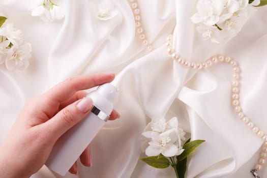 A woman's hand holds a bottle of cream on a background of flowers on a white satin fabric.