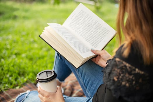 A woman sits near a tree in the park and holds a book and a cup with a hot drink in her hands. A woman in jeans and a t-shirt reading a book outdoor.