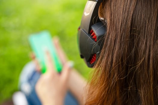 A woman is holding a smartphone and listening to music on headphones while sitting on the grass in a park.