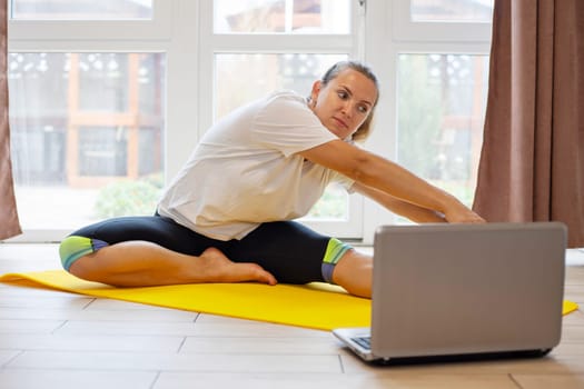 Beautiful young woman in sportswear doing sport exercises on yoga mat at home. Fitness training online with a laptop. Healthy lifestyle concept.
