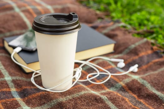 Cup of coffee, open book and smartphone with headphones on blanket in the park or garden.