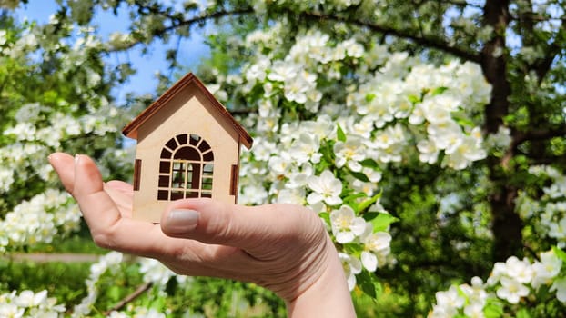 Small wooden toy house on palm of woman hand on natural background with apple flowers. symbol and concept of care, buying, selling, donating of eco friendly home. close-up. soft selective focus