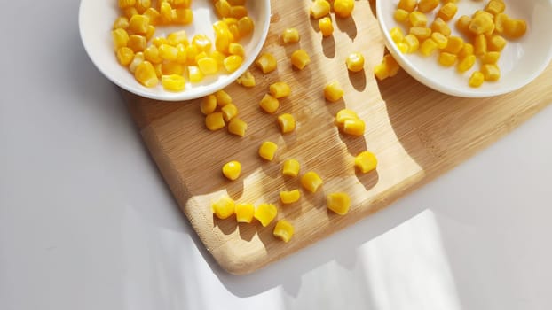 Lot of pieces of canned yellow corn on plate which is on wooden bamboo cutting board on white background. Concept of cooking and delicious healthy food