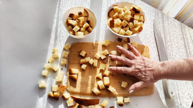 Homemade cooking and cutting of slices of diced dry white rye bread on cutting board and white plate. Hand of woman with Crackers for food, healthy eating on table in sunny day. Top view