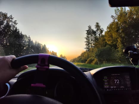view from a car windshield of natural landscape with road, green treeand and light, glare from sun at sunset in evening. Hand of woman on the steering wheel. Female traveler driving on trip or journey