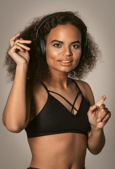 Dancing in headphones young African-American girl listening music wearing black top and make salsa moves with hands in the air, isolated on grey background, emotionally move. 