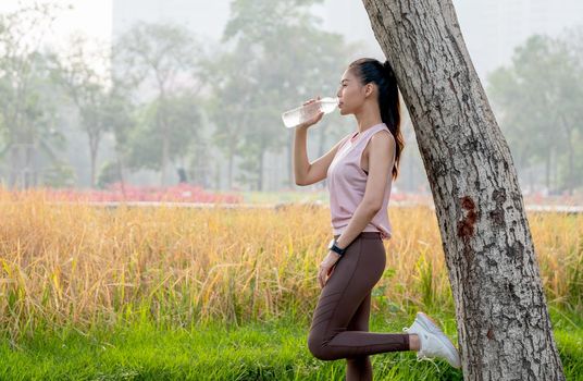 Asian sport woman lean tree and drink water from bottle with grass field as background during exercise in park or garden with morning light.
