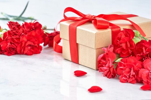 Mother's Day greeting design concept - Beautiful blooming red carnations, gift box with ribbon on white marble table background, close up, copy space.