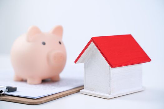 Piggy bank, beautiful wooden house model over a signed contract on white background, concept of saving money to buy a home insurance, close up, copy space.