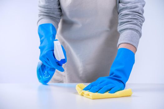 Young woman housekeeper is doing cleaning white table in apron with blue gloves, spray cleaner, wet yellow rag, close up, copy space, blank design concept.