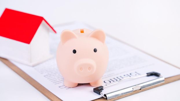 Piggy bank, beautiful wooden house model over a signed contract on white background, concept of saving money to buy a home insurance, close up, copy space.