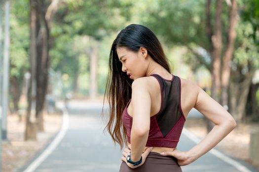 Back of Asian sport woman show action of waist or back pain after exercise in park or garden with day light.
