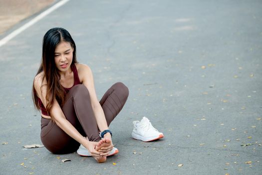 Asian woman with sportswear action of foot pain and sit on road of park or garden during exercise in morning.
