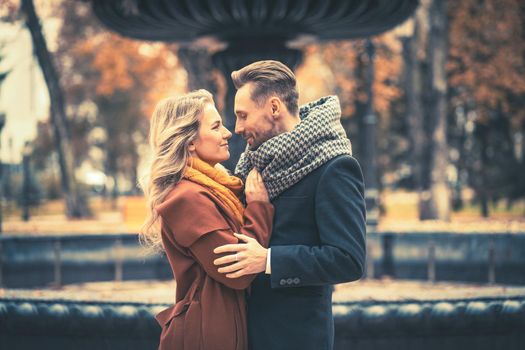 Close up. Loving couple of young people standing embracing and looking at each other near a old fountain in the autumn park wearing autumn coats. Toned photo. 