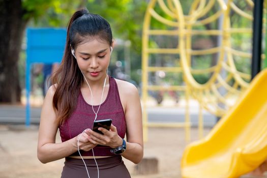 Portrait of Asian woman concentrate to use mobile phone and stay near playground during exercise in park or garden with morning light.