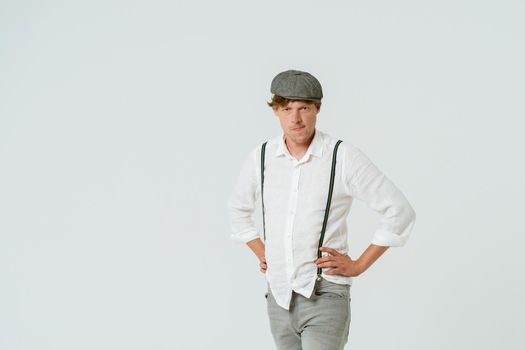 Handsome hipster man posing with hands on hips dressed in white shirt and grey jeans and wool cap isolated on white background. Copy space at left side.