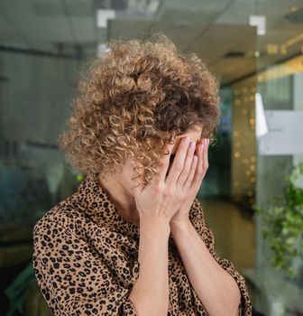 Curly young woman with sad expression covering face with hands while crying in office