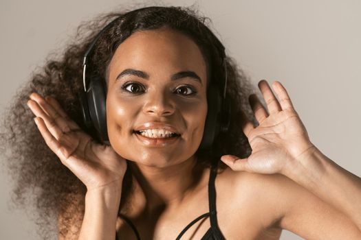 Close up young African-American girl listening music wearing black top in headphones, isolated on grey background, emotionally move, have fun. 