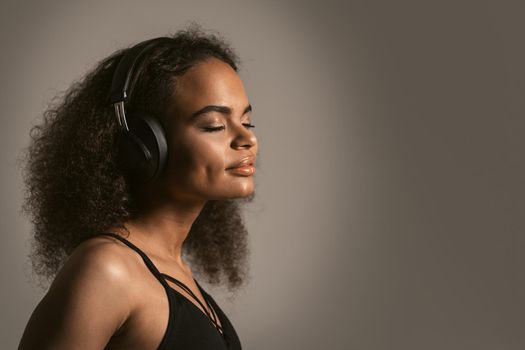 Inspired by music young African-American girl listening music in headphones wearing black top isolated on grey background, emotionally move, have fun. 