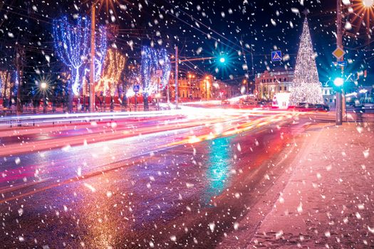 Night city with Christmas decorations, spruce and traces of headlights of moving cars, reflected in the wet asphalt in a snowfall.