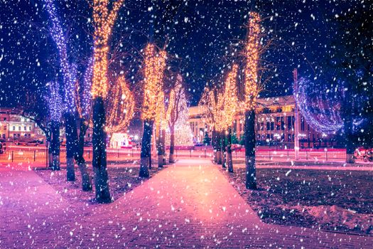 Snowfall in a winter park at night with christmas decorations, lights, pavement covered with snow and trees with garlands.