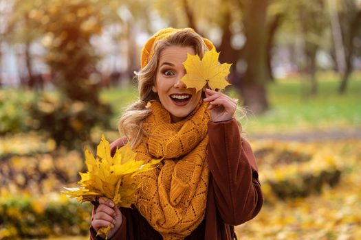 Cheerful young woman looking thru yellow fallen leaf in knitted beret with autumn leaves in hand and fall yellow garden or park. Beautiful smiling young woman in autumn foliage. 