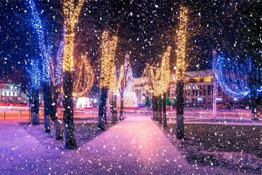 Snowfall in a winter park at night with christmas decorations, lights, pavement covered with snow and trees with garlands.