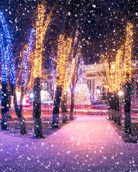 Snowfall in a winter park at night with christmas decorations, lights, pavement covered with snow and trees with garlands.