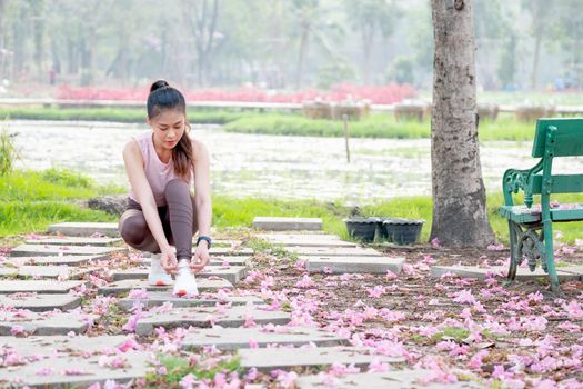 Wide shot of Asian sport woman tie shoelaces on walkway with flower on floor in part or garden during morning.
