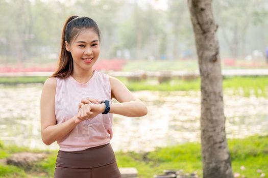 Asian woman with smiling touch her watch and look at camera with lake and flower field as background during her exercise in the morning.