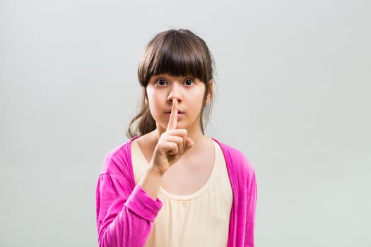 Photo of cute little girl with finger on her lips on grey background.