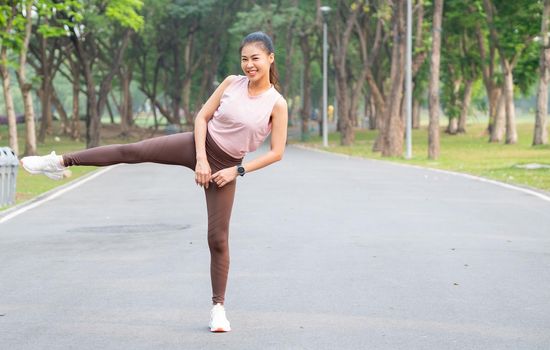 Asian sport woman stand with action of stretch right leg to side also look at camera and smile on road of park or garden with morning light.