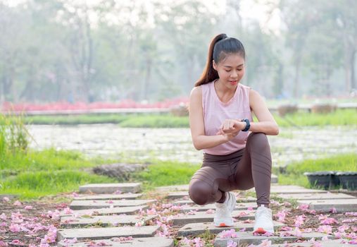 Asian woman with sportwear for jogging sit and look at her watch during exercise in garden or park in the morning with soft light.