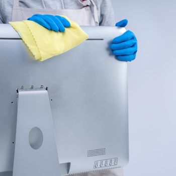 Young woman housekeeper in apron is doing cleaning silver computer screen with blue gloves, wet yellow rag, close up, copy space, blank design concept.