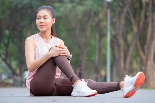 Asian woman with pink sportswear sit with one's knees up and look to her right side on road of park or garden during exercise in the morning.