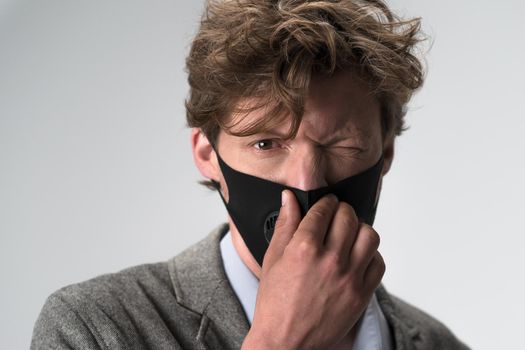 Messy hair handsome young man in grey jacket and a modern reusable protective mask on his face feels a foul smell with using his right hand to cover nose. Epidemic concept. 