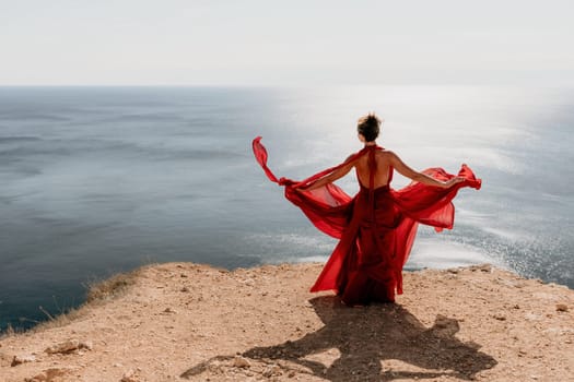 Side view a Young beautiful sensual woman in a red long dress posing on a rock high above the sea during sunrise. Girl on the nature on blue sky background. Fashion photo.