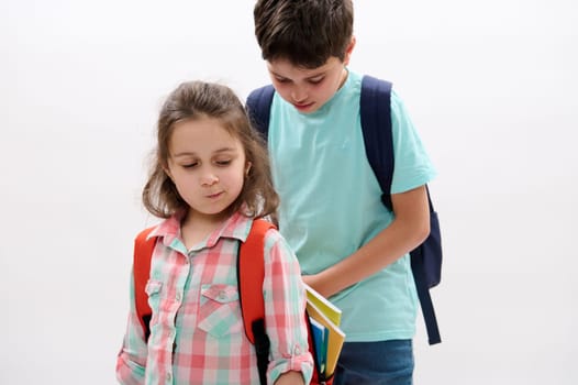 Authentic portrait of a little child girl, first grader or preschooler kid making faces, standing near her older brother, a preteen boy preparing backpack of his sister, over white studio background