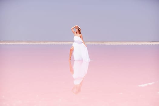 Woman in pink salt lake. She in a white dress and hat enjoys the scenic view of a pink salt lake as she walks along the white, salty shore, creating a lasting memory