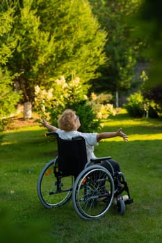 Rear view of an elderly woman spread her arms to the sides while sitting in a wheelchair on a walk outdoors