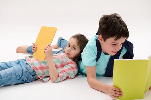 Two charming school kids with backpacks, lying on a white background, holding textbooks, doing homework, resting after school day. September. Education. Back to school on new semester of academic year