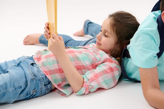 Little girl 6 years old, first grader, primary school student in plaid shirt and blue jeans, holding textbook, lying on white studio background, near her older brother. School kid. Education. Learning