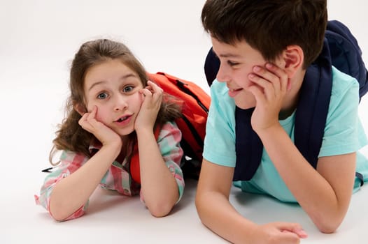 Two adorable smart school kids, a lovely first grader little child girl and her caring brother - a preteen schoolboy, lying on belly, smiling talking relaxing in recreation time over white background