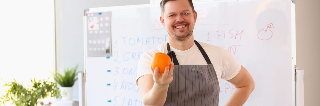 Male cook teacher in apron holds an orange and fresh vegetables and fruits on table. Culinary training and dietary nutrition