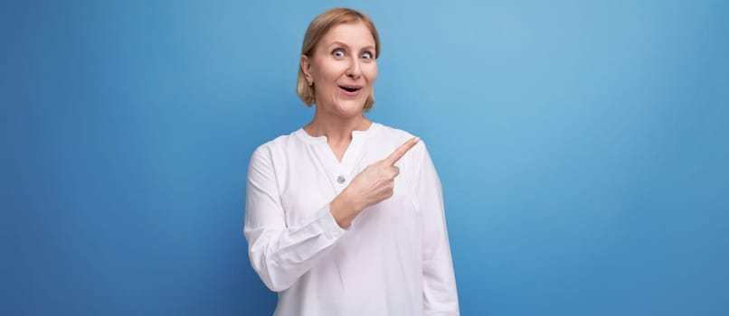 confident blond middle aged woman in white blouse pointing her finger to the side on studio background with copyspace.